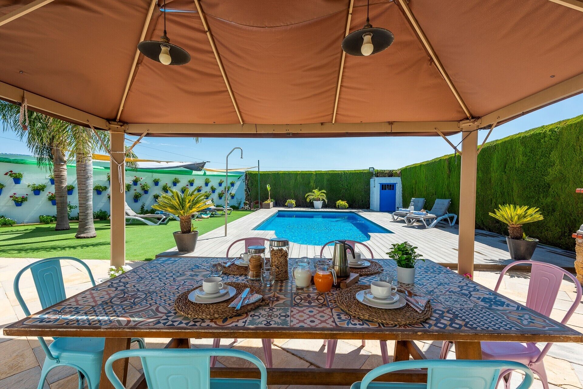 An outdoor patio with a pool, a covered dining area, and a blue wall with potted plants.