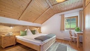 A bedroom with a wooden ceiling, a bed with pillows, a nightstand, a window with a view, and a chair.