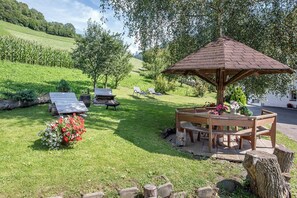 Un kiosque avec un banc et une table en bois, entouré d’une pelouse agrémentée d’arbres et offrant une vue sur un champ.
