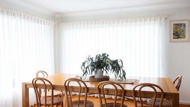 Uma sala de jantar com mesa e cadeiras de madeira, um vaso com plantas e cortinas leves.