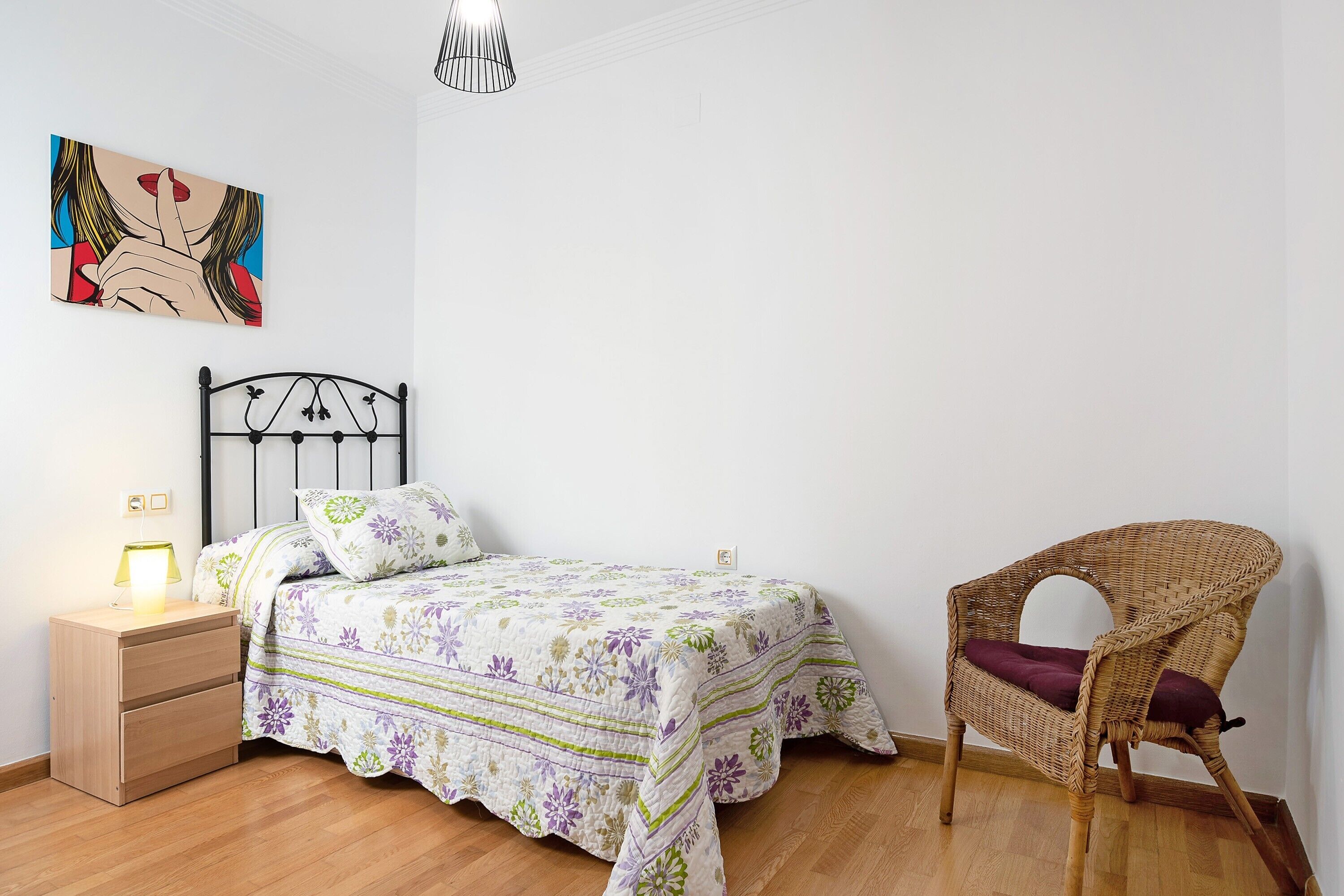 A bedroom with a bed, a nightstand, a chair, a wall art piece, and a hanging light fixture.
