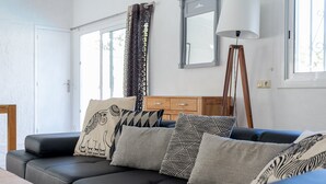 A modern living room with a black leather sofa, patterned cushions, a wooden cabinet, and a floor lamp. - Villa 'Sea près des golfs-El Sol Naciente' with Mountain View, Pool and Wi-Fi (Santa Cristina d'Aro)