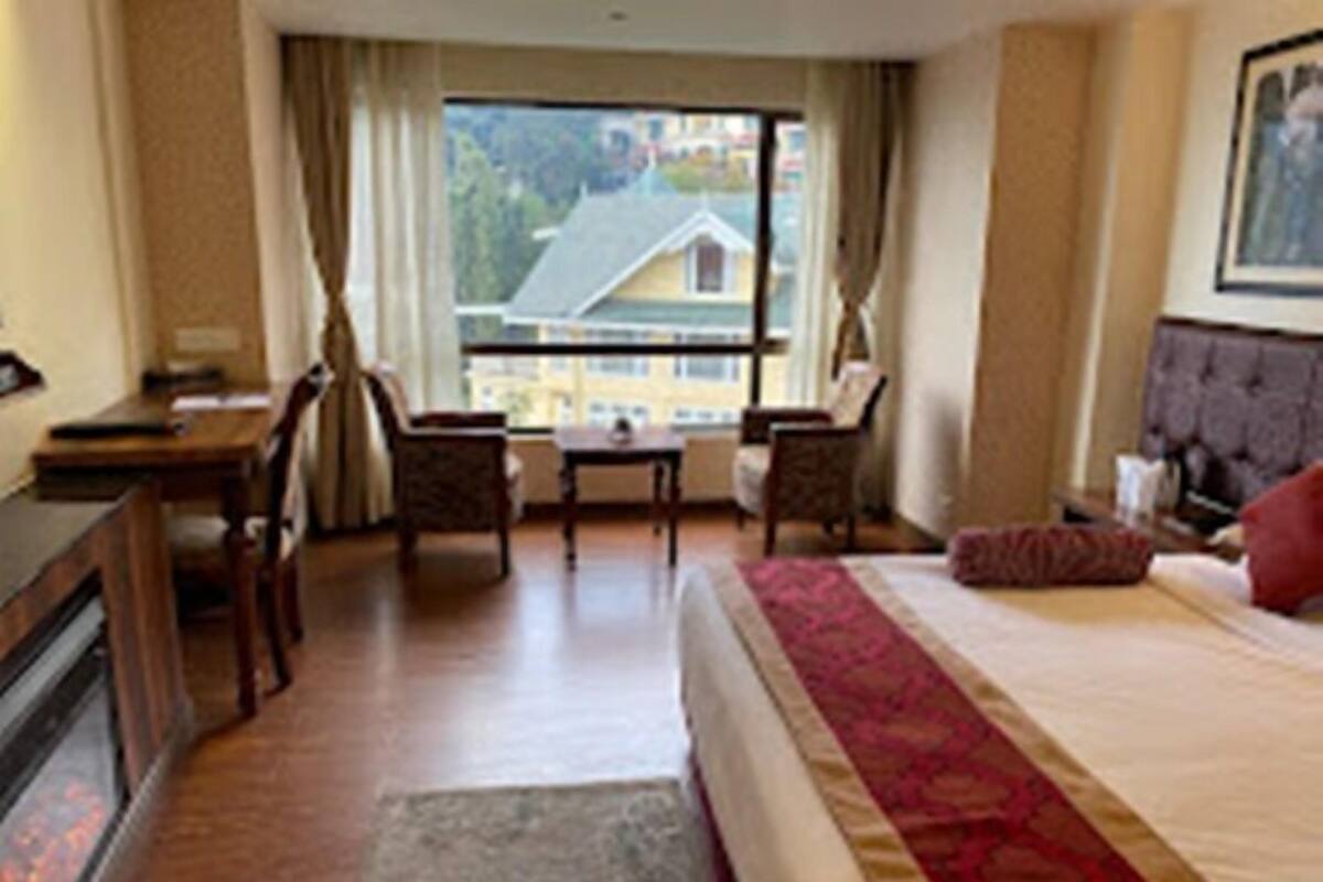 A hotel room with a bed, a desk, two chairs, a window with a view of buildings, and a fireplace.