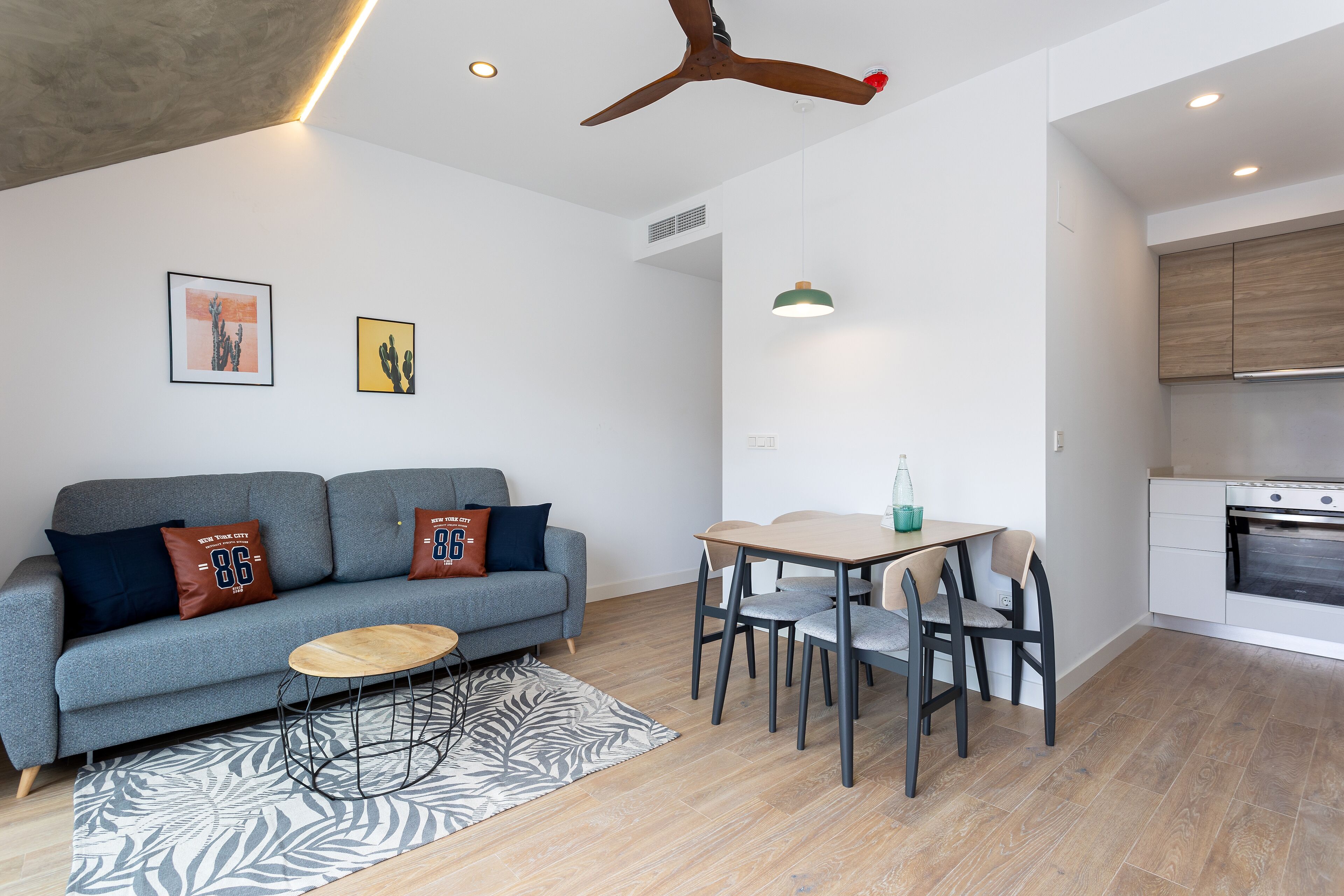 A modern living room with a grey sofa, a wooden coffee table, and a dining area with a table and chairs. There is a kitchen area with a stove and cabinets.