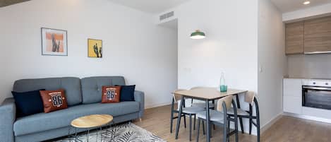 A modern living room with a grey sofa, a wooden coffee table, and a dining area with a table and chairs. There is a kitchen area with a stove and cabinets.