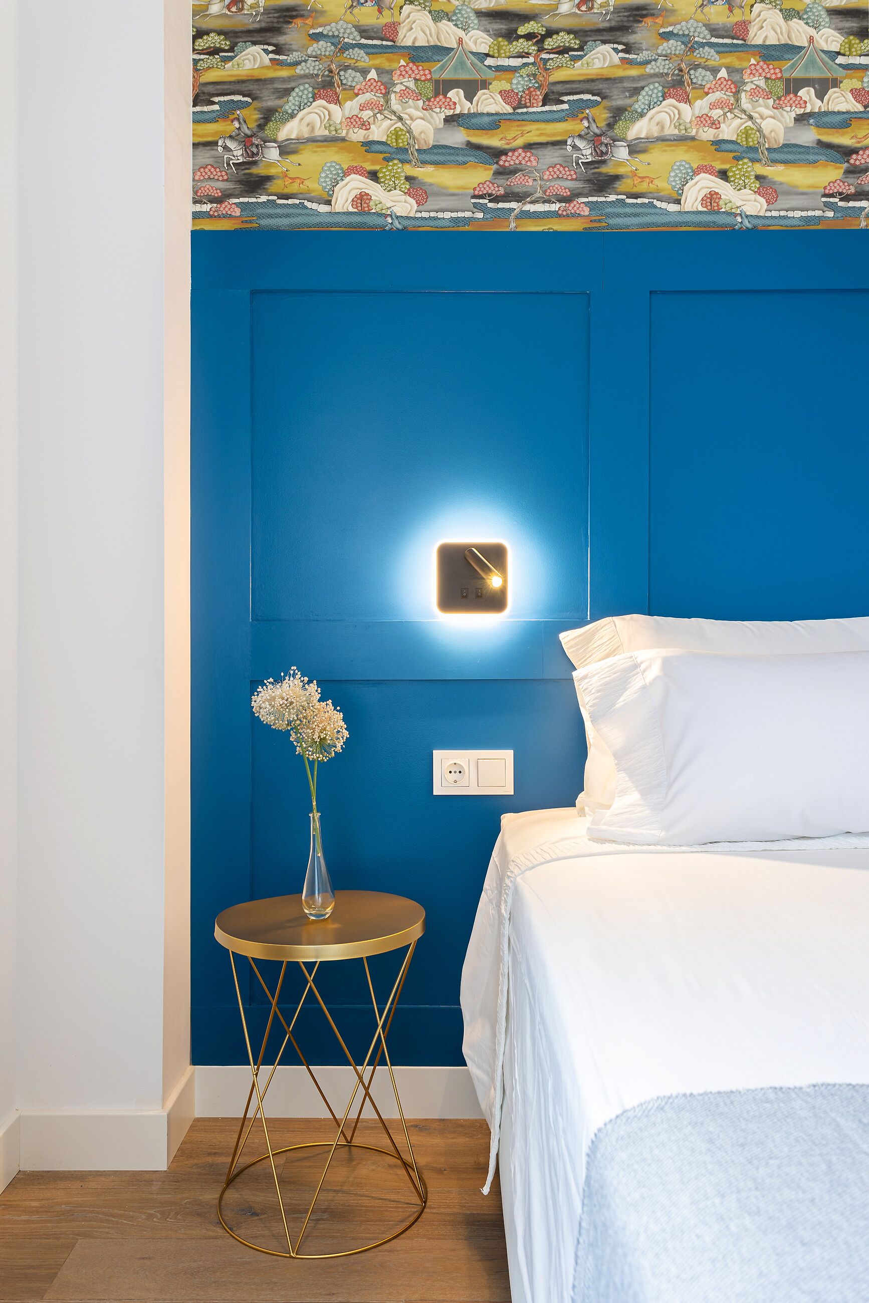 Quarto com parede azul de destaque, cama com roupa de cama branca, uma pequena mesa redonda com um vaso de flores e uma luminária fixada na parede.
