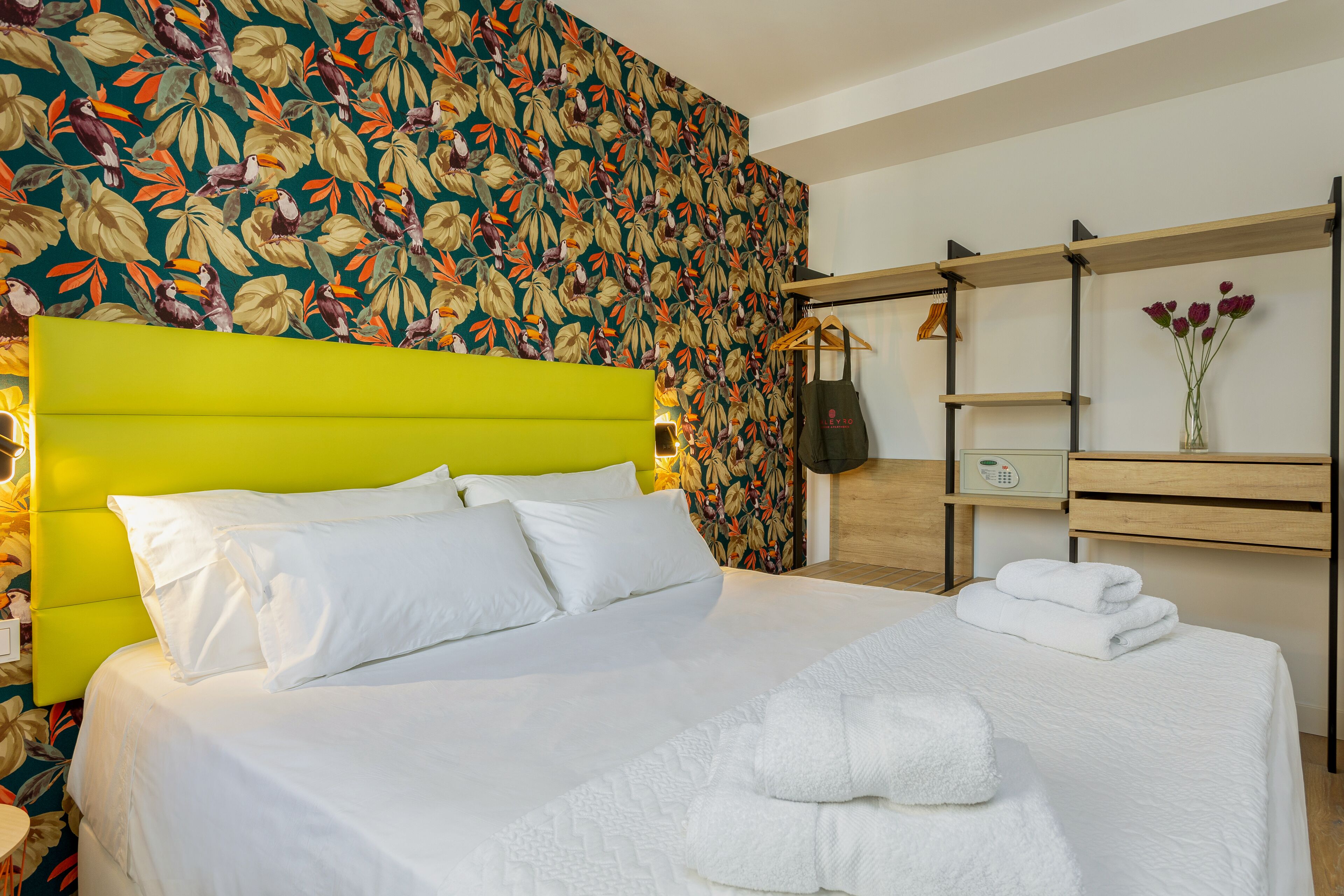 A hotel room with a bed, a bright green headboard, a floral wallpaper, a wooden nightstand, a shelf with a vase of flowers, and a hanging bag.