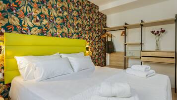 A hotel room with a bed, a bright green headboard, a floral wallpaper, a wooden nightstand, a shelf with a vase of flowers, and a hanging bag.