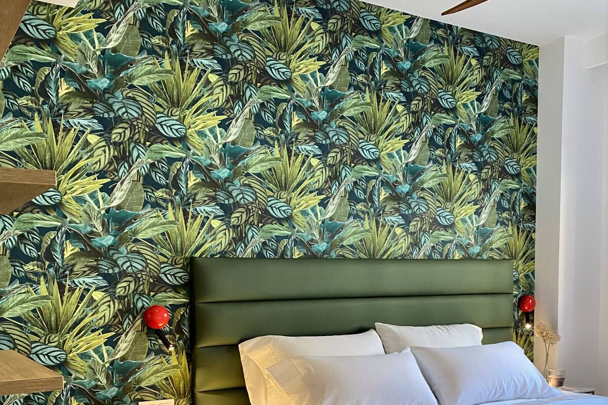 A bedroom with a bed, a nightstand, a lamp, and a wall with a tropical leaf pattern.