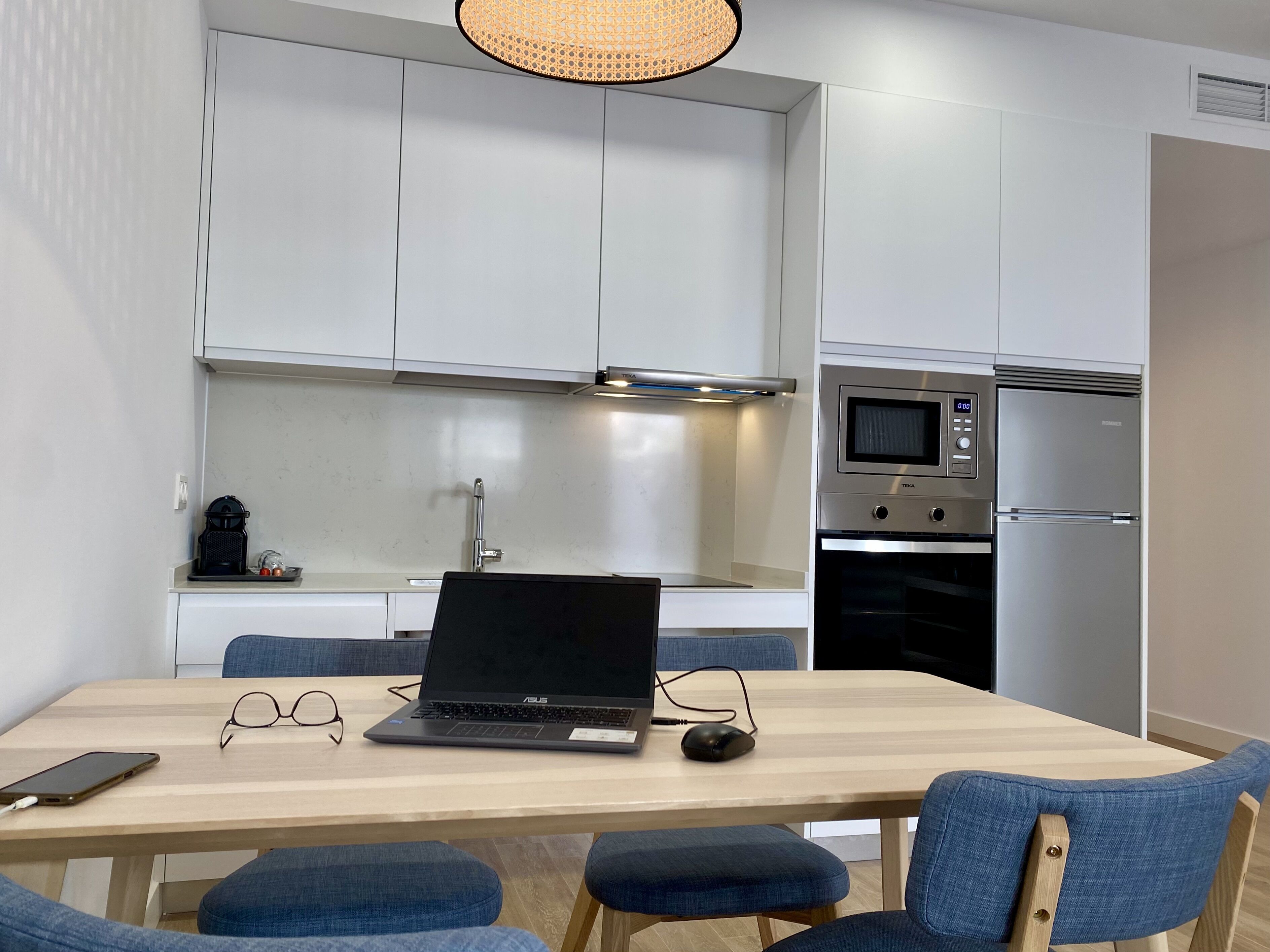 Eine moderne Küche mit weißen Schränken, einer eingebauten Mikrowelle und einem Backofen. Auf einem Holztisch steht ein Laptop, daneben Gläser und eine Maus.