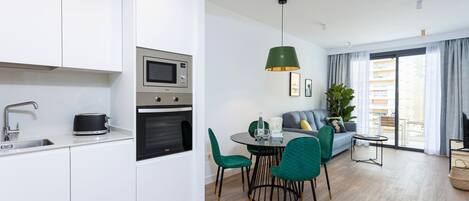 A modern kitchen with white cabinets, a microwave, and a stove. There is a dining area with a round table, green chairs, and a hanging light fixture.