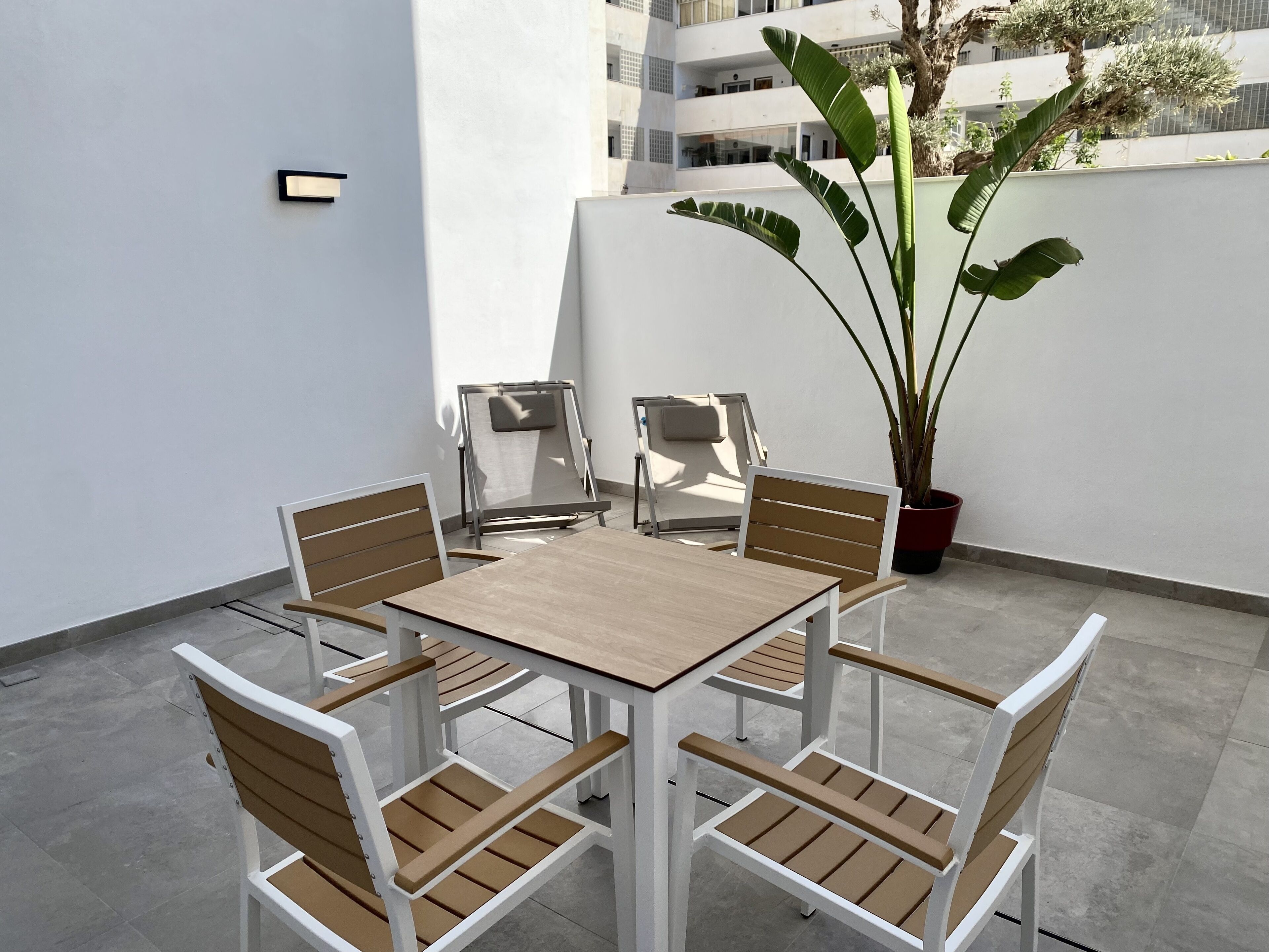 Eine moderne Terrassenanlage mit einem quadratischen Tisch, sechs Stühlen, einem großen Blumentopf und einer weißen Wand.