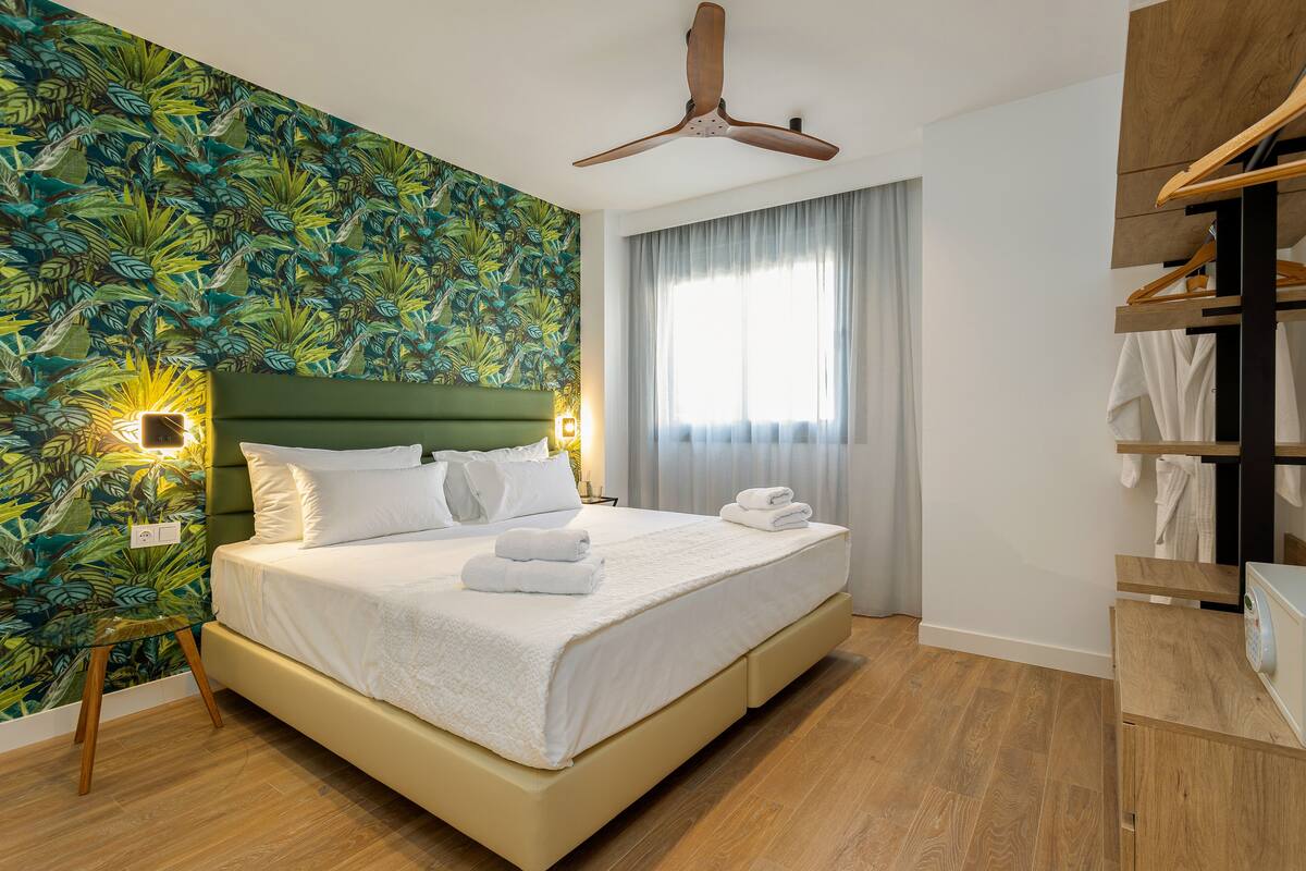 A neatly made bed with white linens and towels, a wooden headboard, and a tropical leaf-patterned wall.