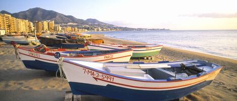 Several boats lined up on a sandy beach with a mountainous backdrop and buildings in the distance.