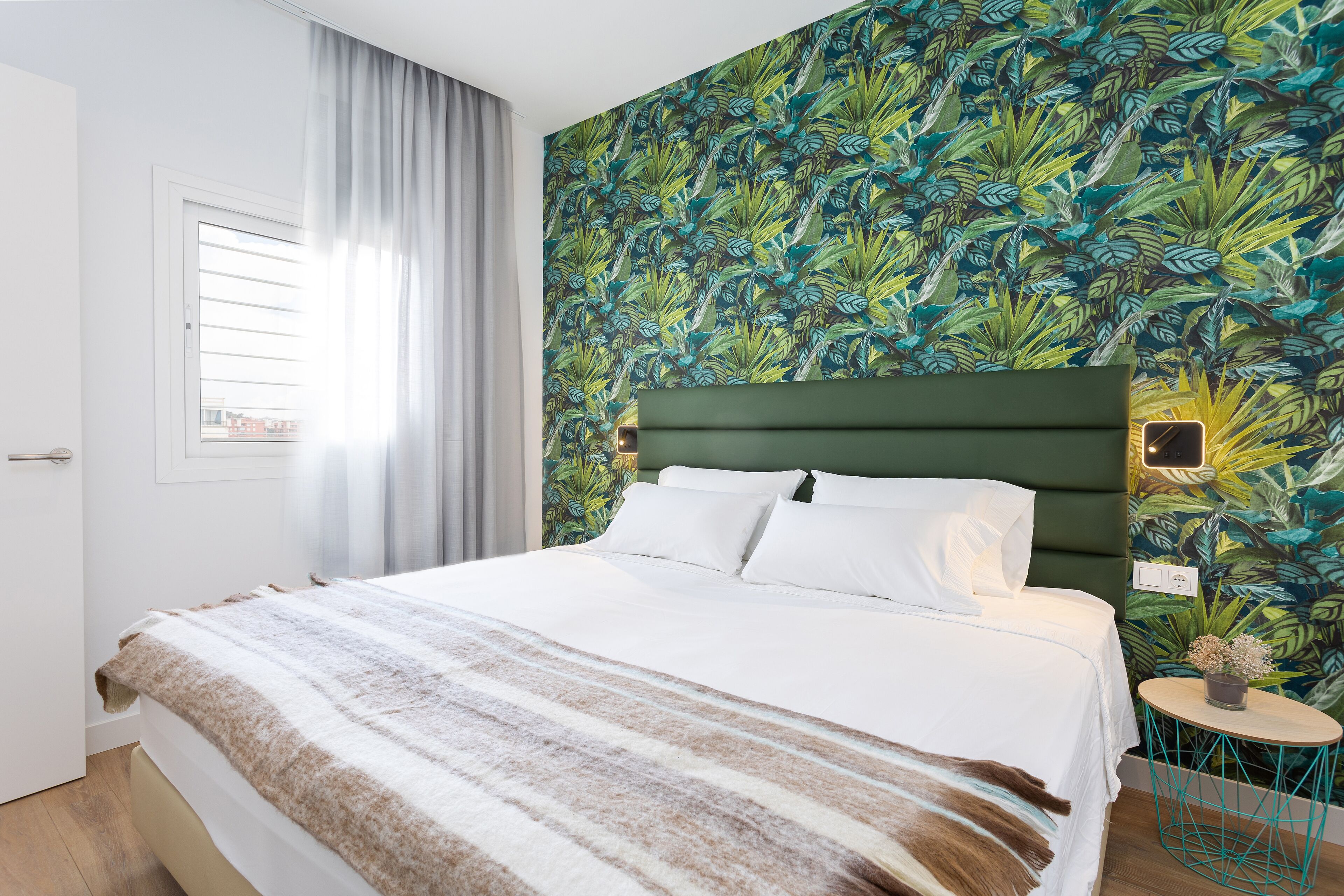 A neatly made bed with a striped blanket, a green headboard, and a tropical leaf-patterned wall.