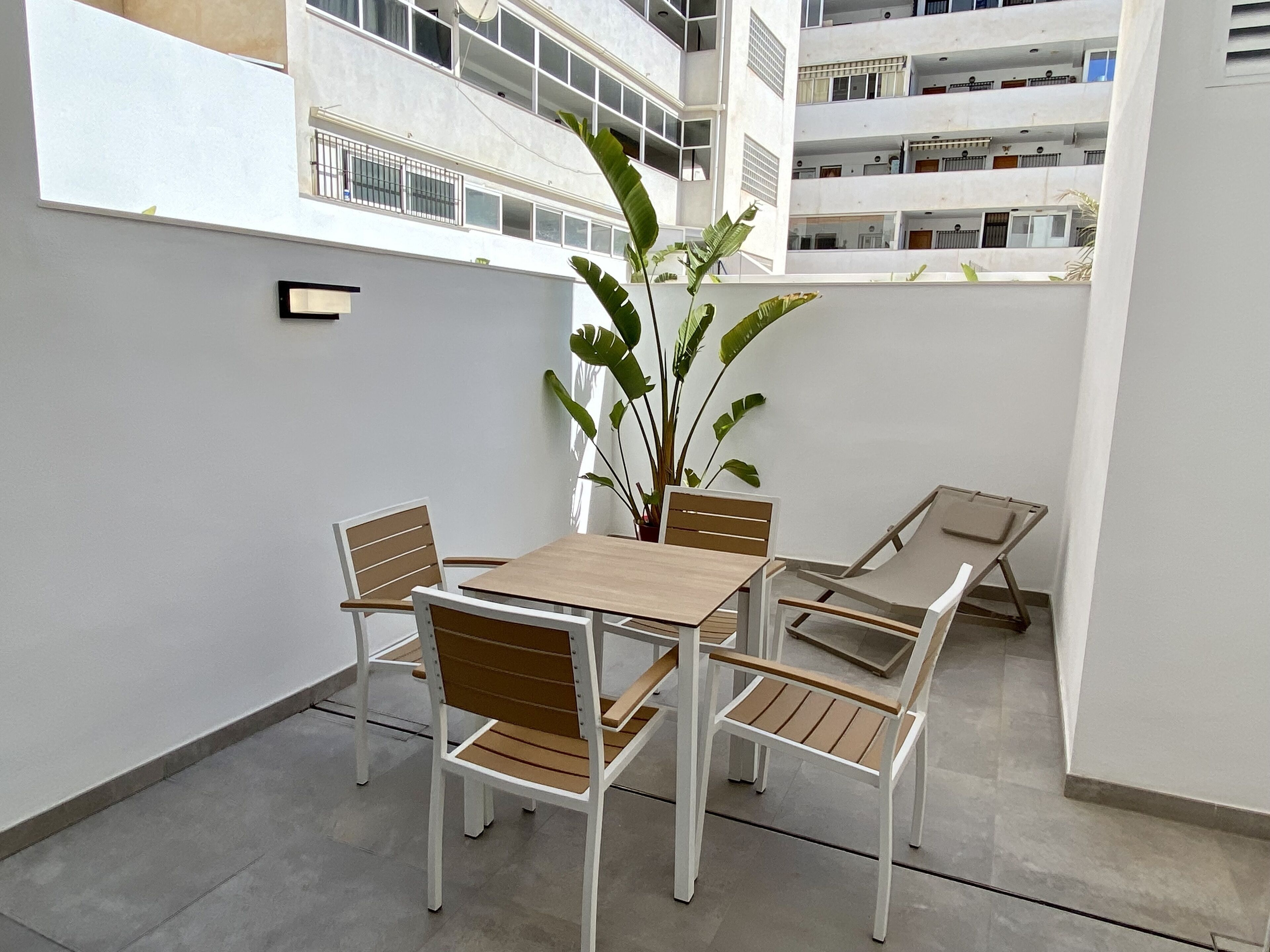 A small outdoor patio with a wooden table and chairs, surrounded by white walls and a large plant.