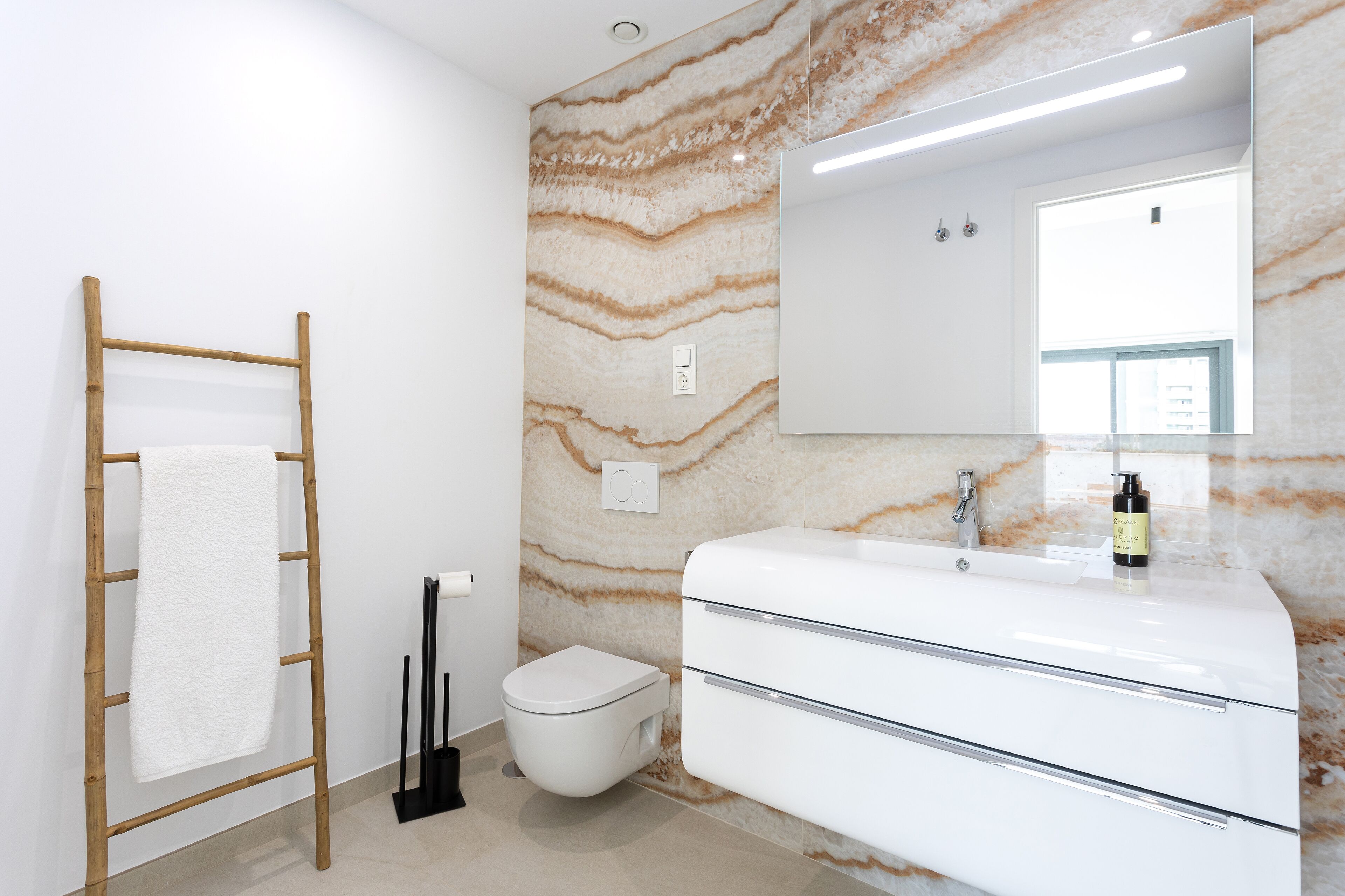 Un baño con pared de mármol, toallero de madera, inodoro y lavamanos blancos, y un espejo sobre el lavamanos.