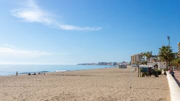 A wide sandy beach with a clear blue sky, a few people, and buildings in the background.