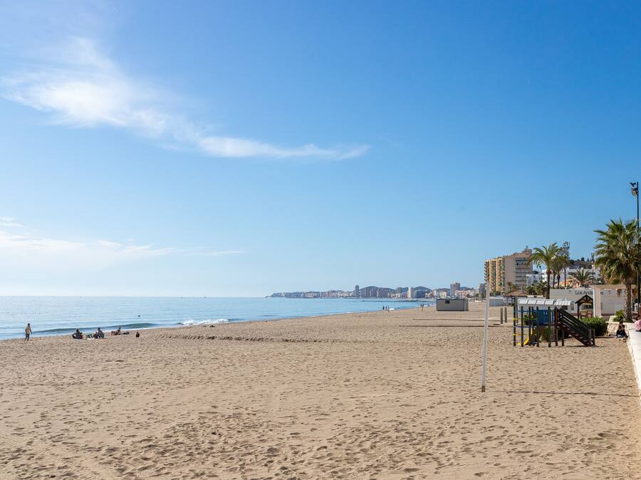 A wide sandy beach with a clear blue sky, a few people, and buildings in the background.