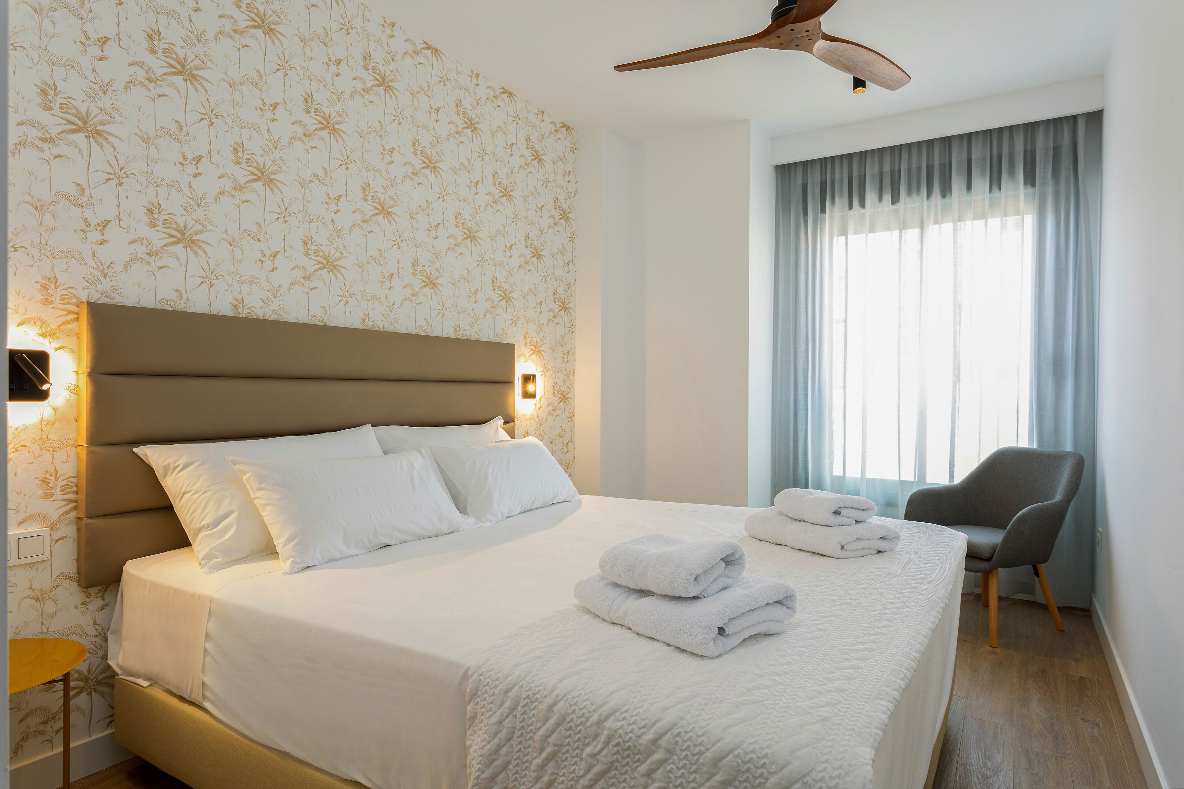 Een net opgemaakt bed met witte lakens en kussens, een nachtkastje met een lamp, een stoel en een raam met gordijnen.