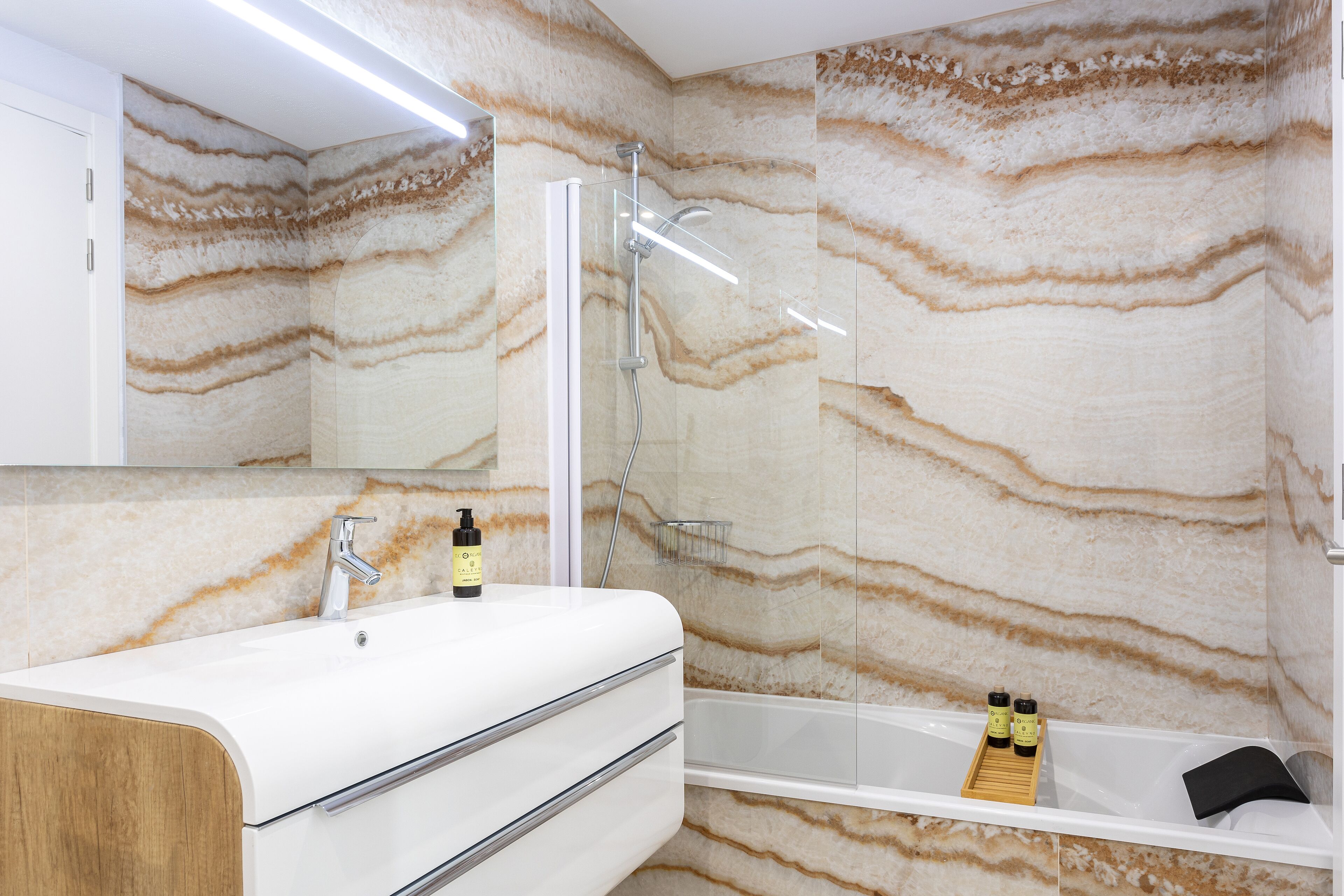 Un baño moderno con pared con diseño de mármol, un lavamanos blanco con gabinete de madera, ducha envuelta en vidrio y una bañera con bandeja de madera.