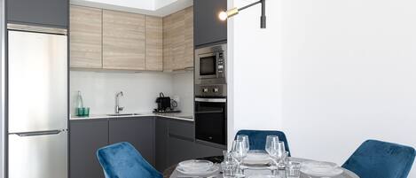 A modern kitchen with grey cabinets, a stainless steel refrigerator, and a microwave. There is a dining table set with blue chairs and clear glassware.
