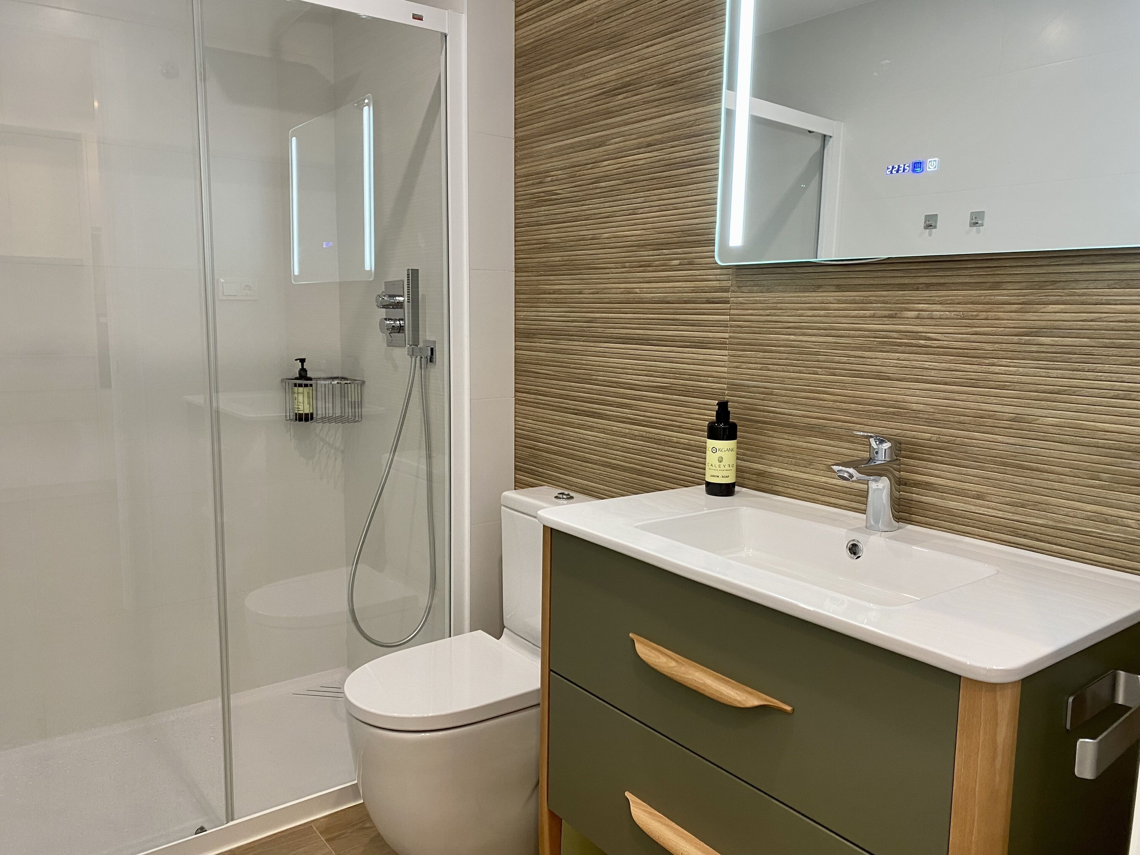 Une salle de bain moderne avec une douche à parois de verre, des toilettes blanches, un meuble-lavabo avec un miroir et une bouteille de shampoing posée sur le lavabo.