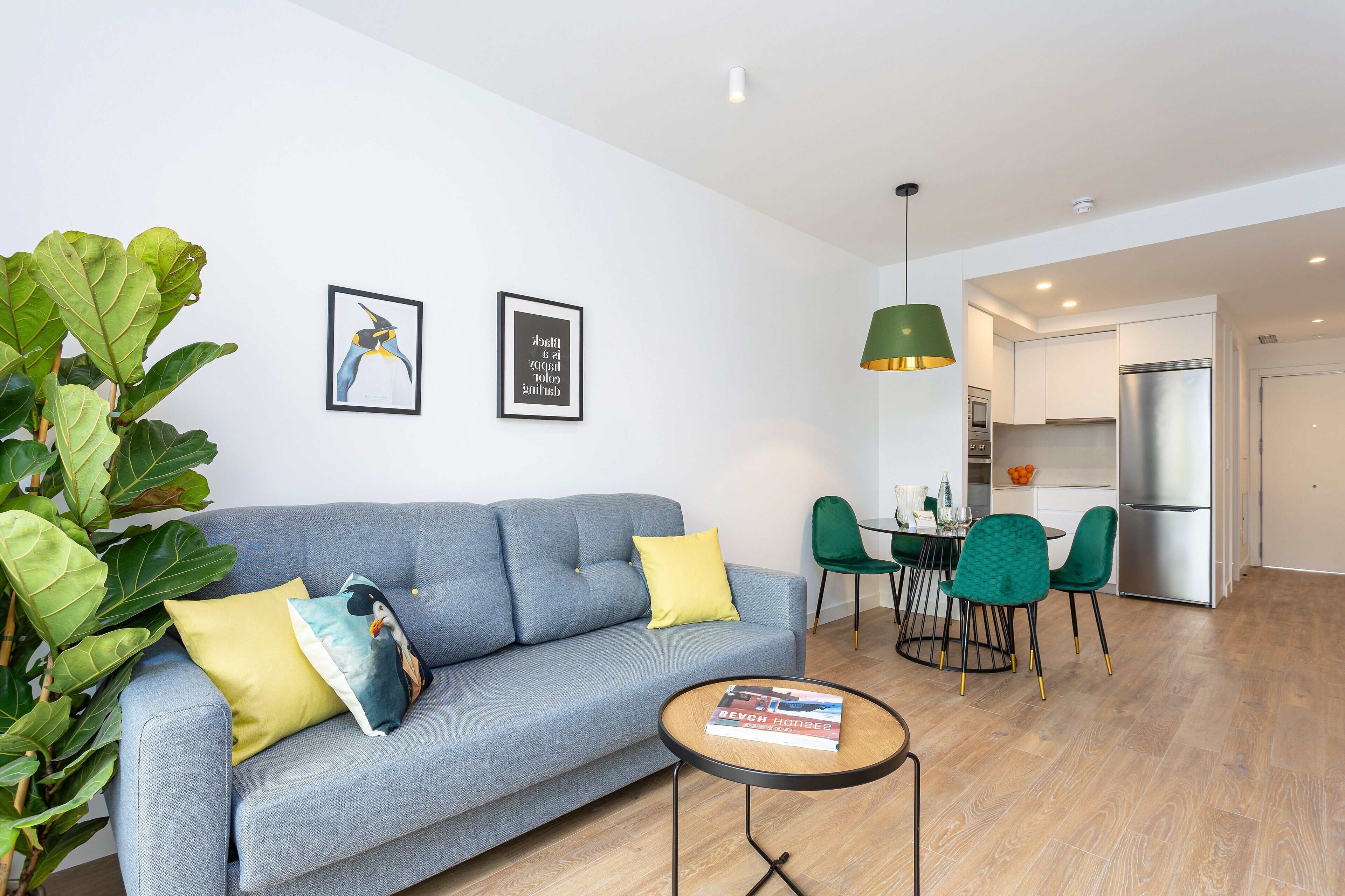 A modern living room with a grey sofa, a small round table, and a dining area with green chairs.