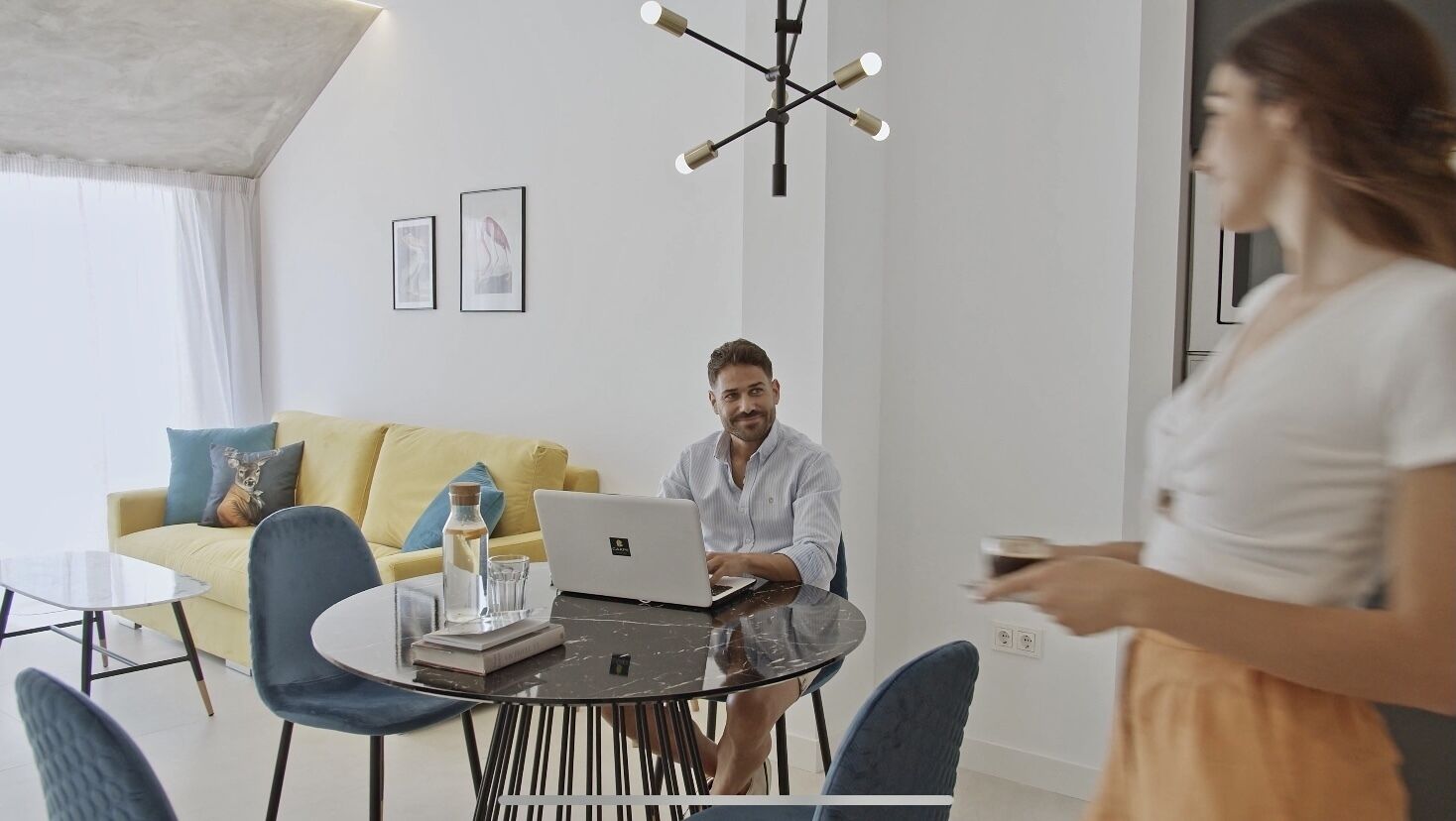 Una sala de estar moderna con un sofá amarillo, una mesa de centro de vidrio y una computadora portátil sobre una mesa redonda.