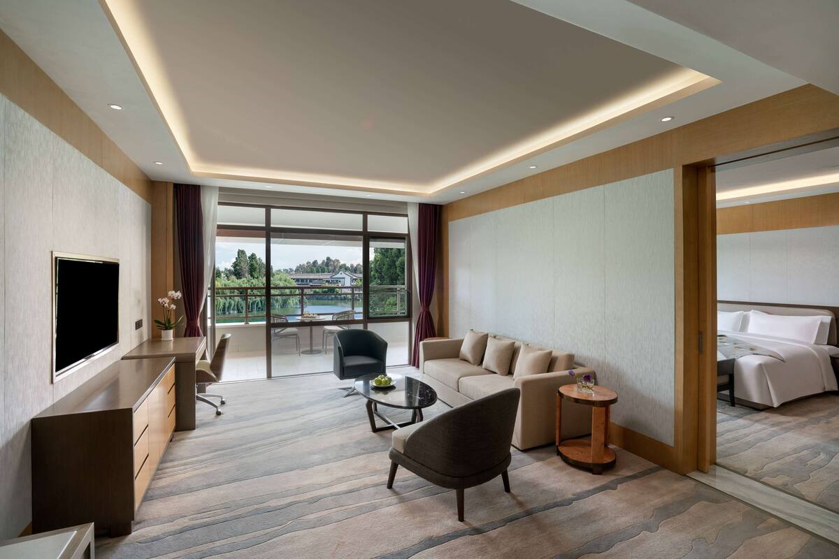 Een moderne hotelkamer met een groot bed, een bank, een bureau met een televisie en een balkon met uitzicht op bomen en een meer.