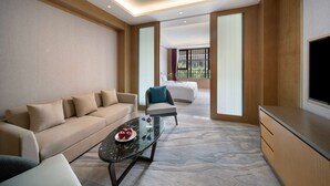 A modern hotel room with a sofa, a coffee table with fruit, a TV, and a large window offering a view of greenery.
