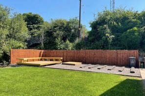Un patio trasero con terraza de madera, zona de barbacoa y jardín cercado.