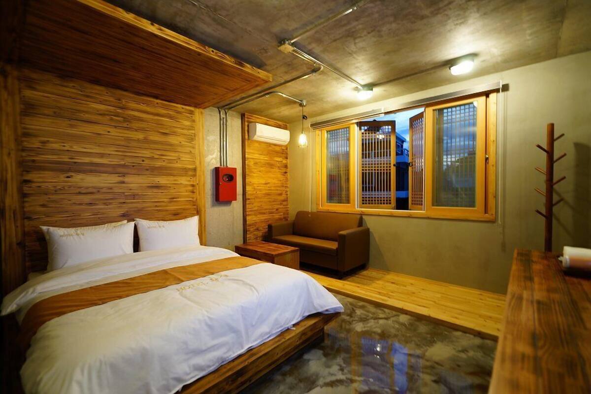 A bedroom with a wooden headboard, a bed with white bedding, a brown sofa, and a window with a view of the outside.