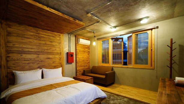 A bedroom with a wooden headboard, a bed with white bedding, a brown sofa, and a window with a view of the outside. - Hwangsil Tourist Hotel (Jeonju)