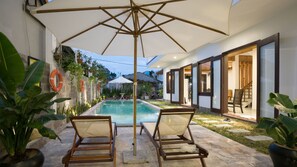 Relax by the pool surrounded by lush greenery and enjoy the shade under a large umbrella.