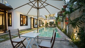 A swimming pool with a table and chairs, surrounded by a garden and a building with large windows. - Hoi An Maison Vui Villa (Da Nang Municipality)