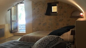 Une chambre confortable avec un mur en pierre, une fenêtre avec des rideaux, un lit agrémenté d’un coussin à motifs et une table de chevet avec une lampe.