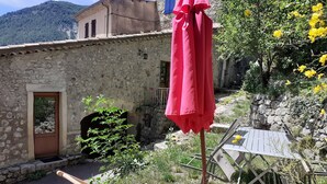 Un espace extérieur rustique agrémenté d’un mur en pierre, d’une porte en bois, d’un parasol rouge et d’une chaise pliante.