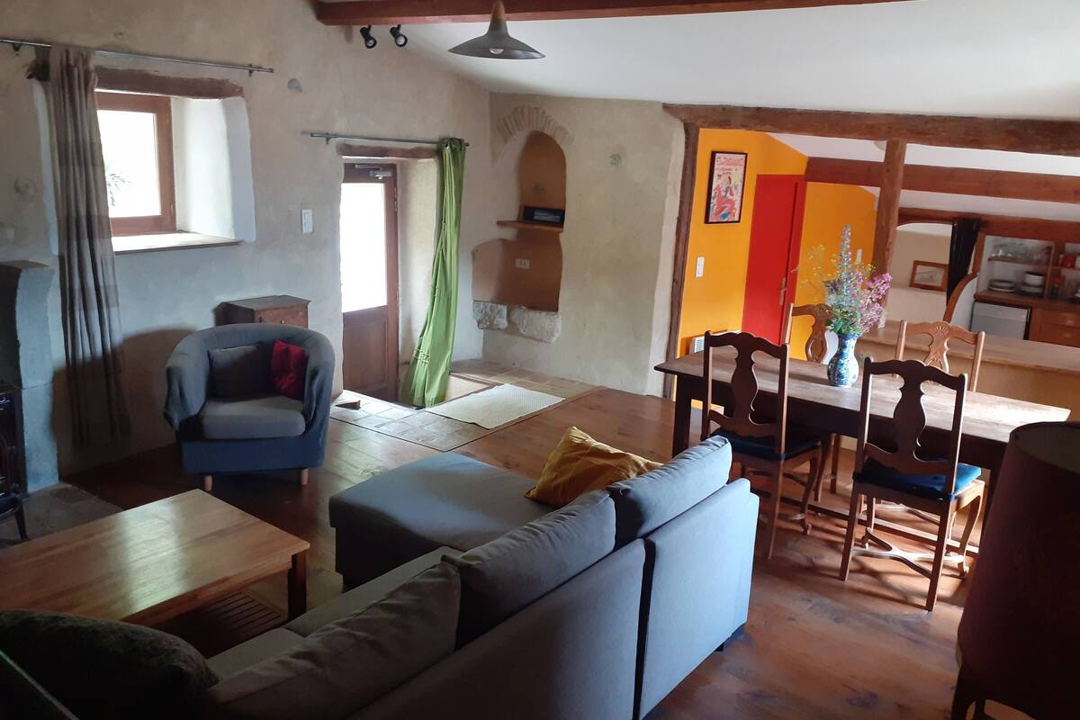 A living room with a sofa, a dining table with chairs, a kitchen area, and a window with curtains.