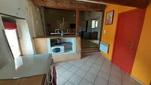Une cuisine équipée d’un four à micro-ondes, d’un évier et d’un petit plan de travail. Une poutre en bois se trouve au-dessus du plan de travail, et une porte rouge est située sur le côté droit.