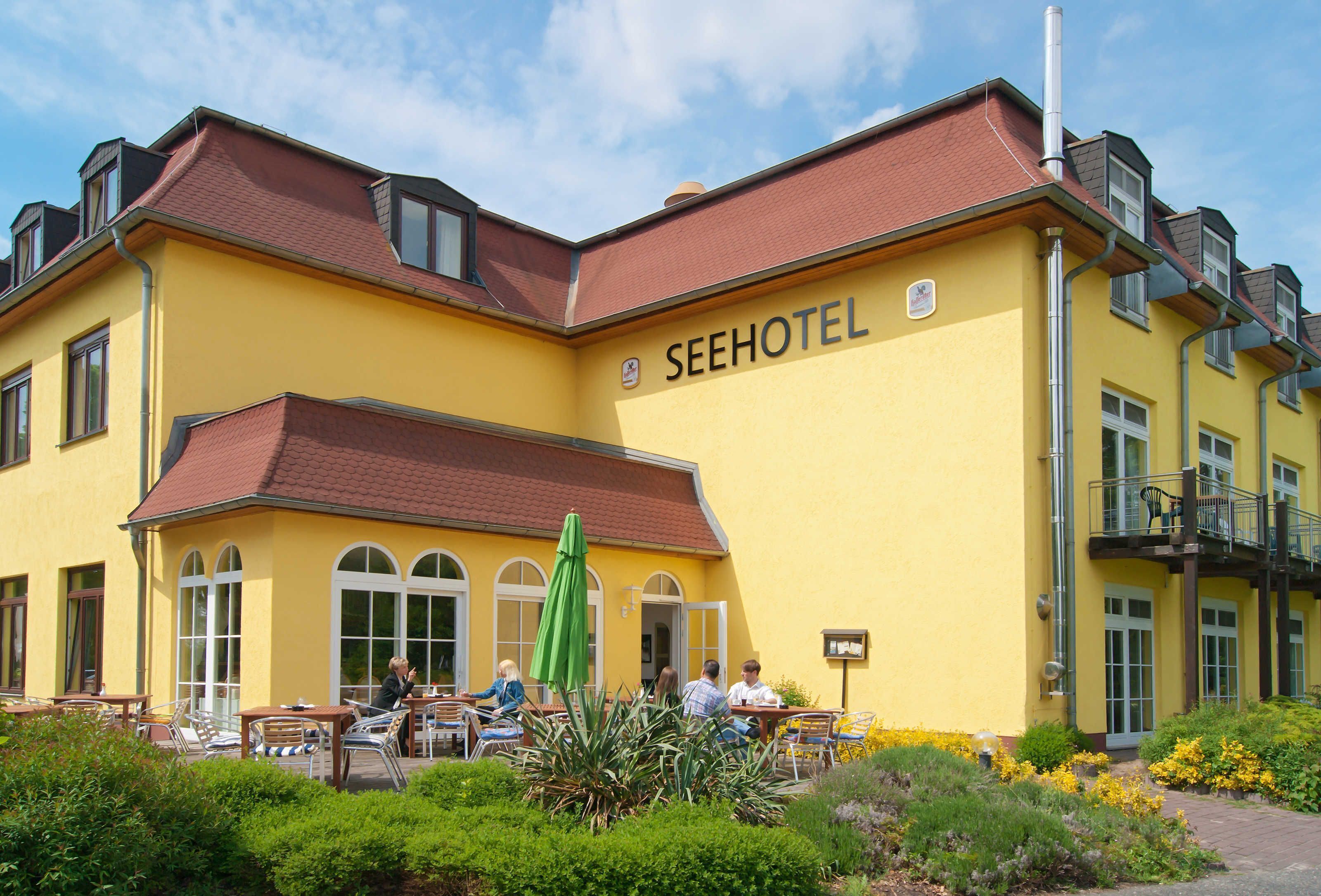 A yellow building with the sign SEEHOTEL, outdoor seating with umbrellas, and a garden area.