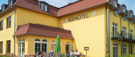 A yellow building with the sign SEEHOTEL, outdoor seating with umbrellas, and a garden area.