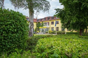 A yellow building with a reception area, surrounded by lush greenery and trees. - Junior suite - Seehotel Brandenburg. (Beetzsee)
