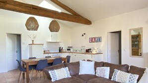 Un salón acogedor con un sofá seccional marrón, una mesa de centro de madera y un comedor con mesa y sillas de madera. Se ve una cocina con armarios blancos y un letrero que dice 'CAFE' al fondo.