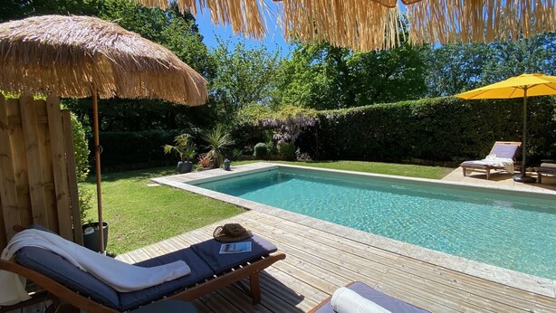 Un área junto a la piscina con sillones de descanso, una sombrilla de paja y una terraza de madera.