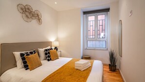 A neatly made bed with a white and mustard blanket, a headboard, and decorative pillows. A small bedside table with a lamp is visible. - Ruby Orange Apartment, WiFi, AC !New! (Lisboa)