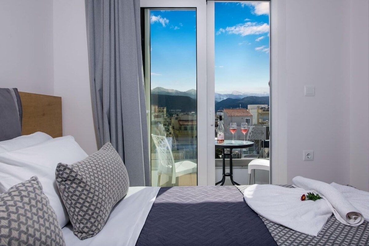 A hotel room with a bed, a small table with two glasses of red wine, and a view of mountains through a glass door.