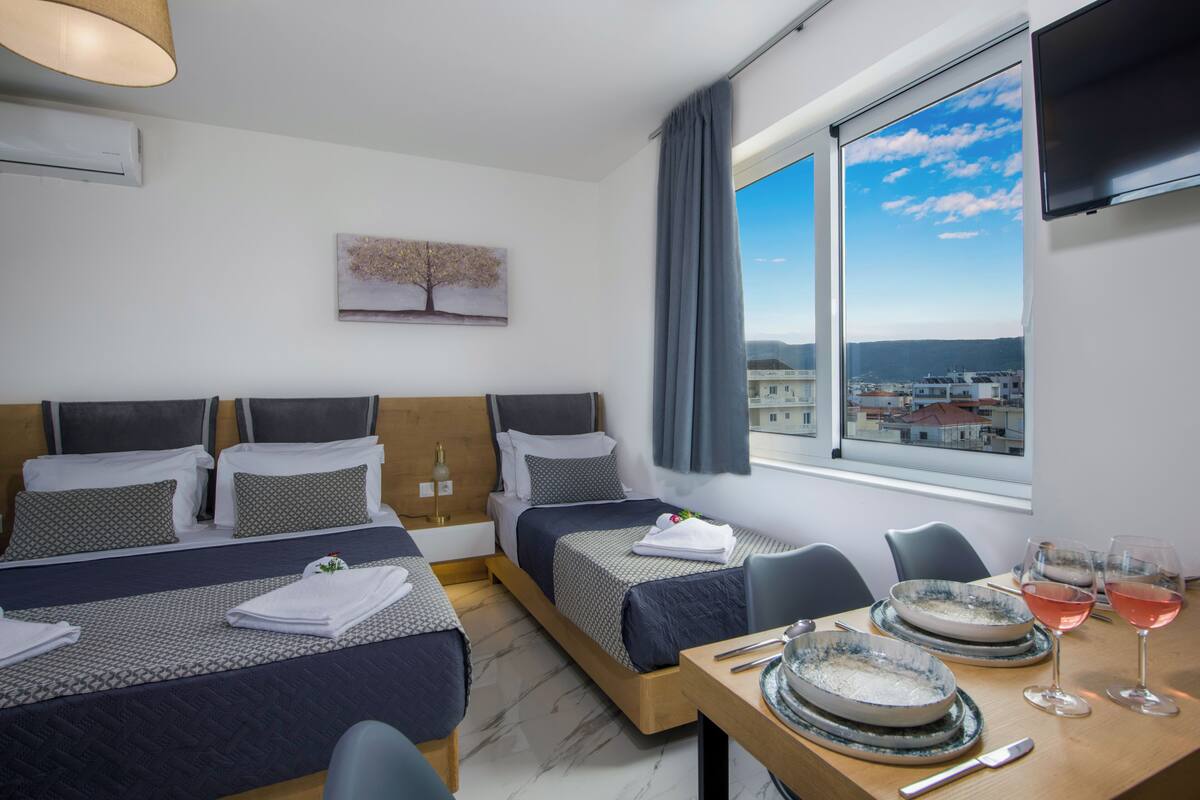 A hotel room with two beds, a dining table with plates and glasses, and a view of the city through large windows.