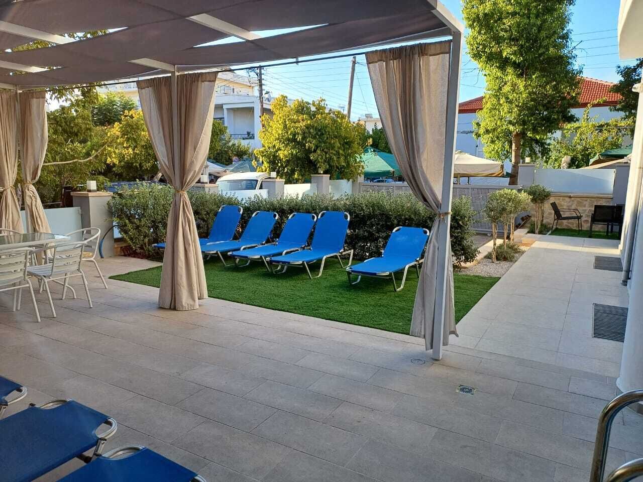 A covered outdoor seating area with blue chairs and a table, surrounded by greenery and a clear sky.