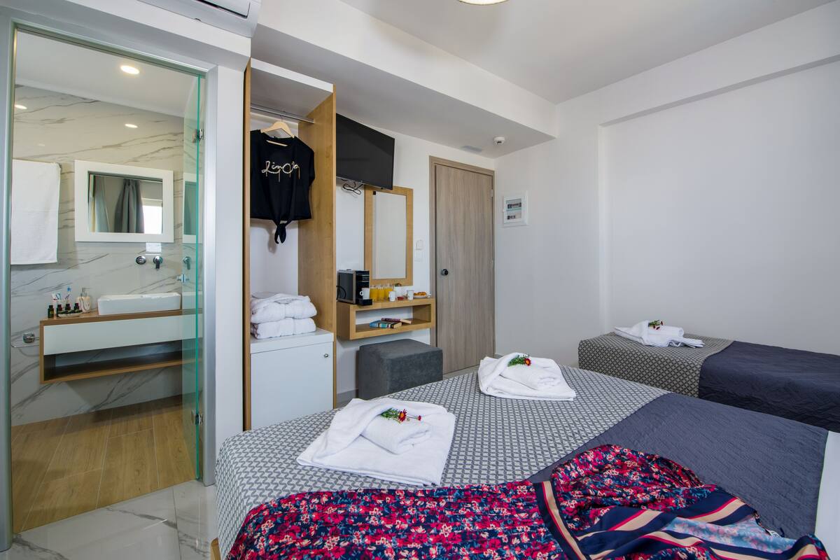 A hotel room with a glass shower enclosure, a bed with a floral bedspread, a bedside table with a lamp, and a wardrobe with a mirror and a television.